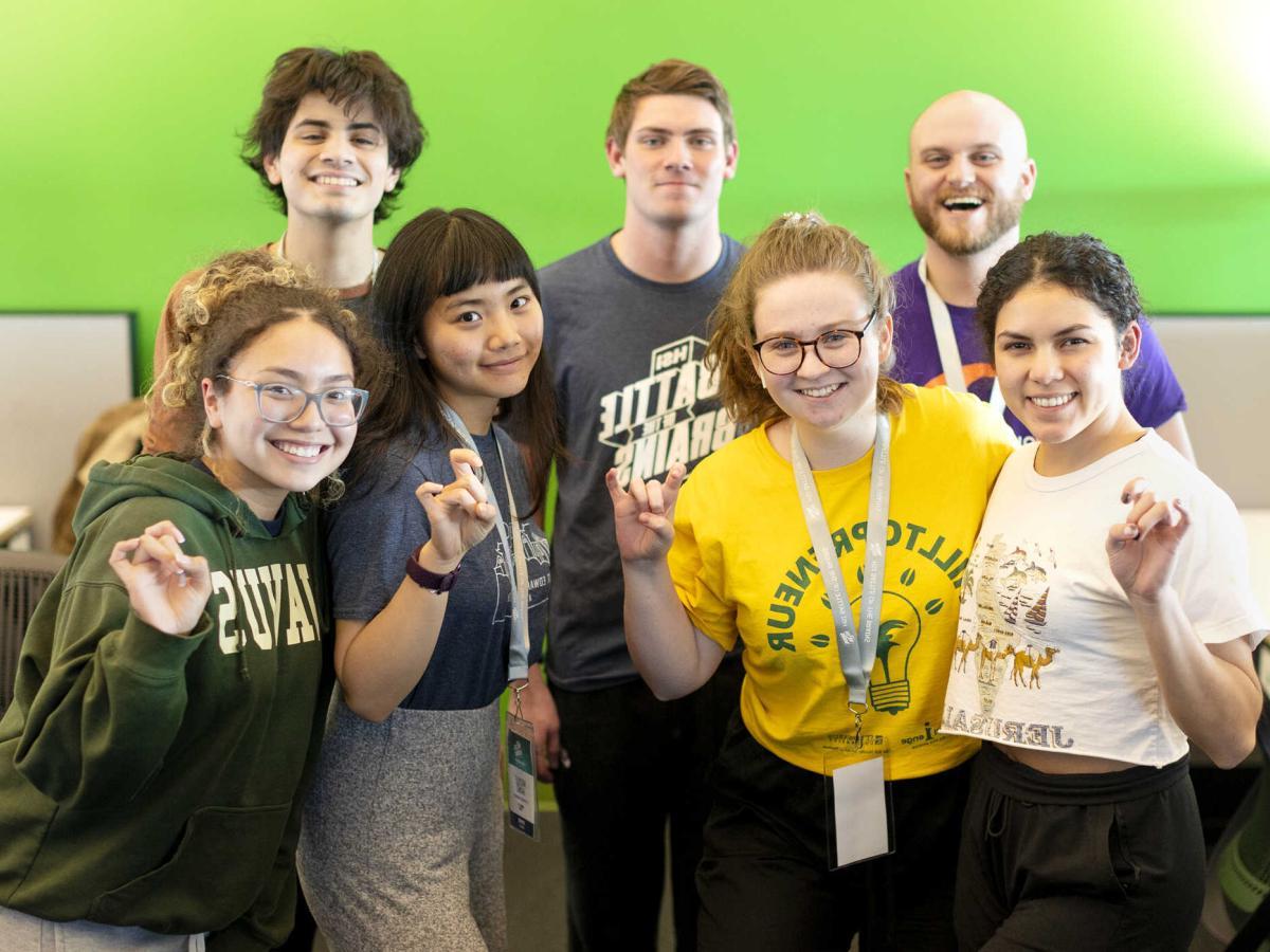 Seven students st和 in front of a green wall 和 give toppers up h和 signs.