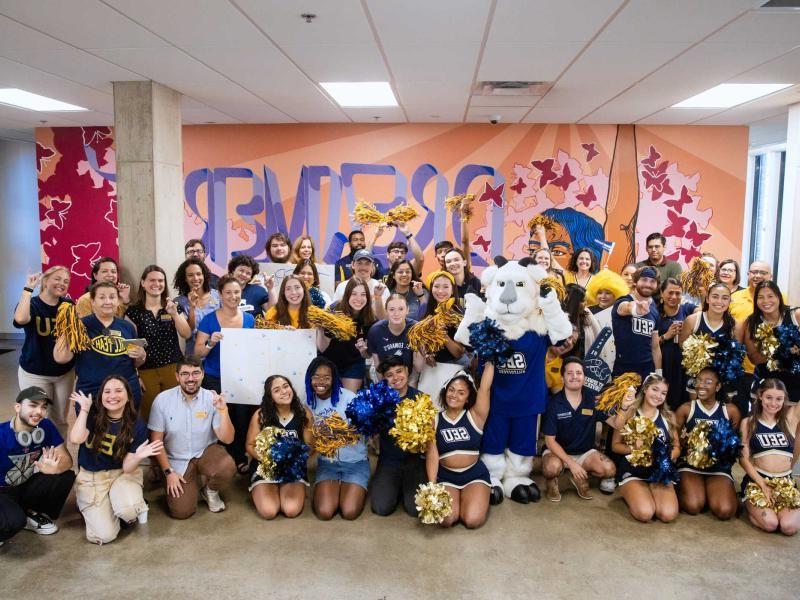 The image depicts a large group of people, including cheerleaders, faculty, and a mascot, posing enthusiastically in front of a colorful mural with the word "DREAMER" prominently displayed. 这群人穿着蓝色和金色的衣服，许多人拿着绒球，其中包括SEU (St. Edward's University) mascot. 气氛是喜庆和庆祝，暗示学校或社区活动.