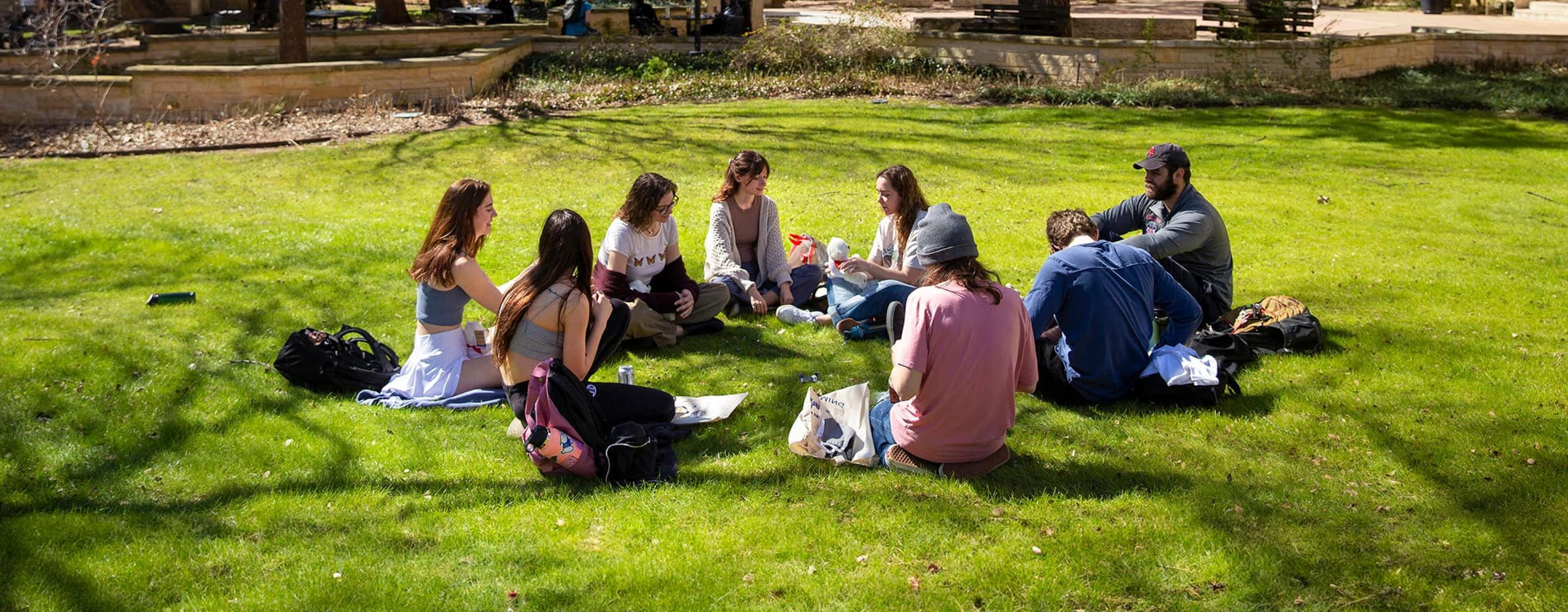 A group of students sitting on the grass in what appears to be a campus setting, with large trees providing shade and buildings in the background. The individuals are arranged in a circle, some with their backs to the camera, and appear to be engaged in a group activity or discussion. The scene is set on a sunny day with clear skies.