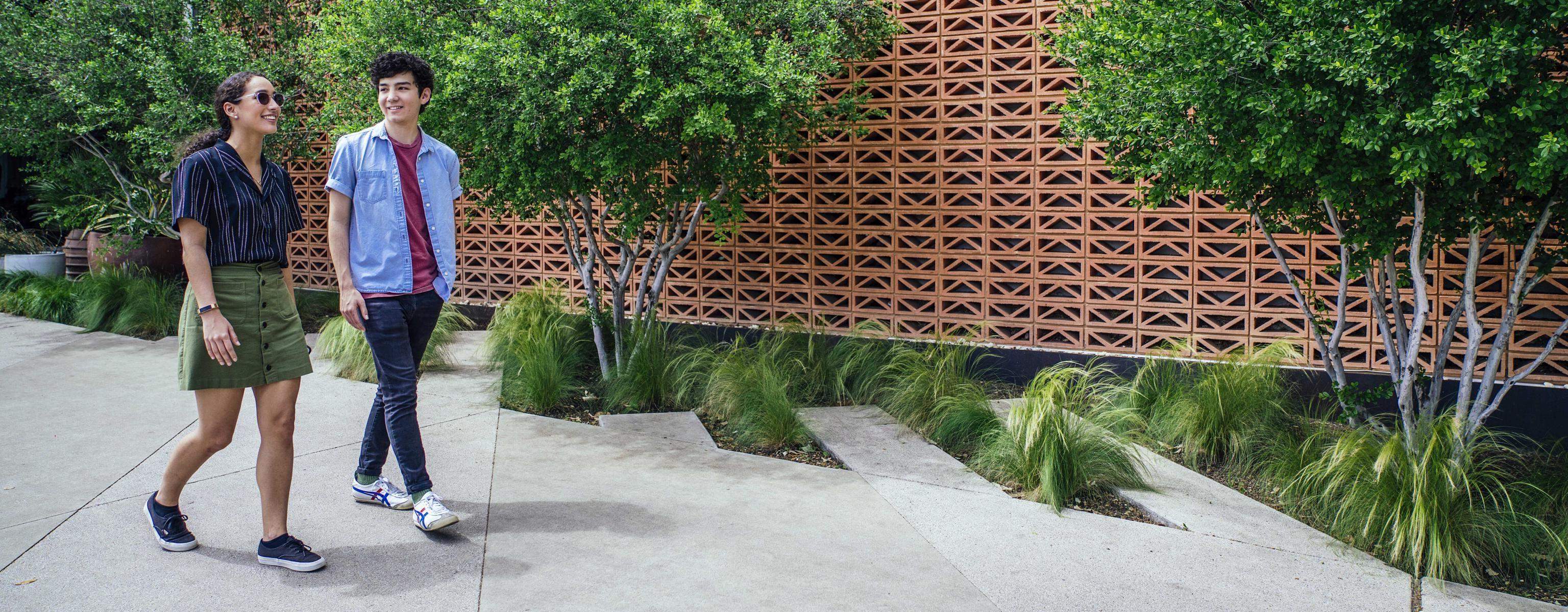 Two students walk by a earth-toned patterned stone wall with trees in front of it on South Congress.