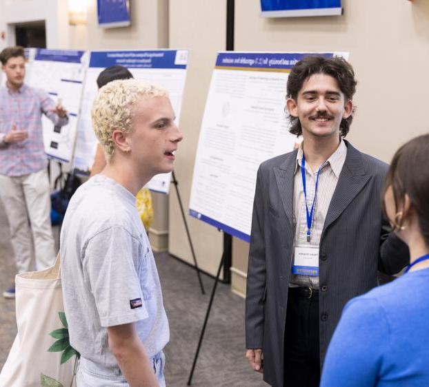 A student presenter talks with other students in front of their research poster.