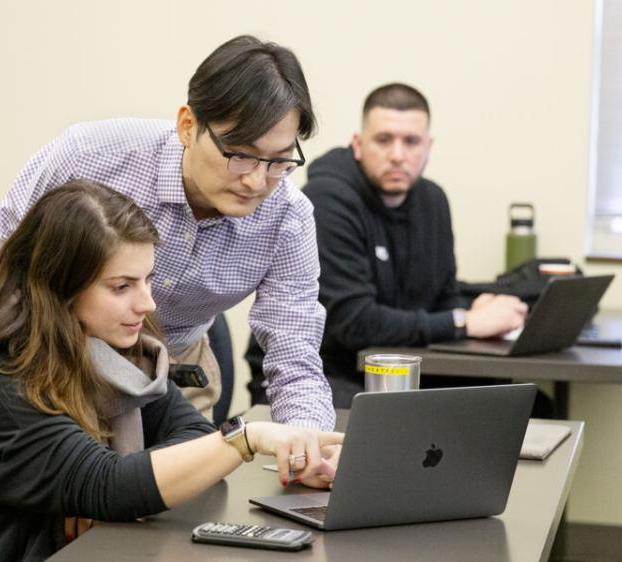 A student watches as a professor assists another student on the student's laptop in class.