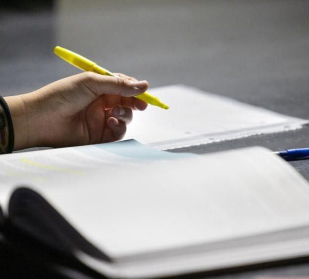 A hand holds a highlighter on top of a notebook at a desk.