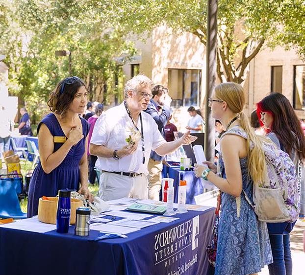 Faculty from the School of Behavioral and Social Sciences stand at a table on Ragsdale Lawn and talk to students.