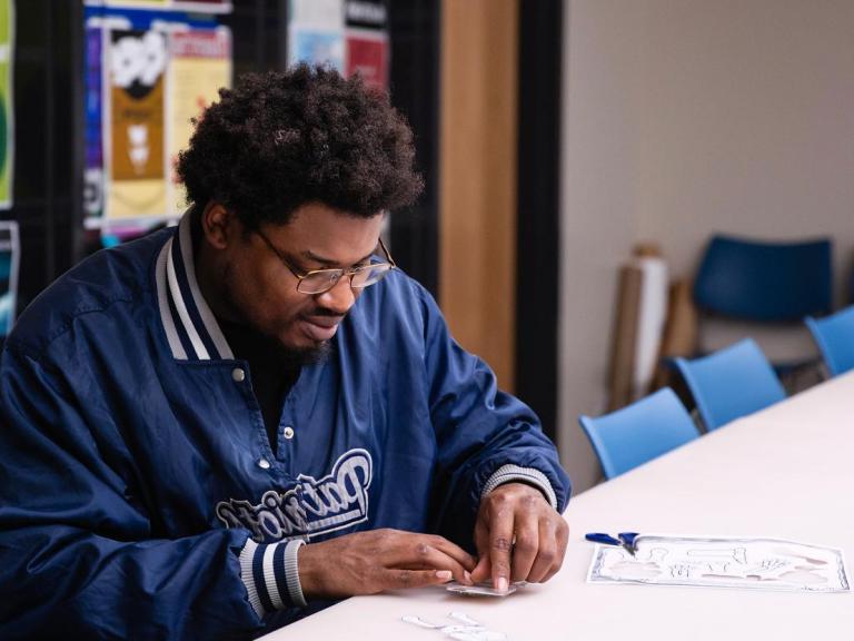 The image shows a man with glasses and a beard, wearing a blue "Patriots" jacket, 坐在桌子旁，专注于一个手工项目，包括剪纸. 他正在一个房间里独自工作，房间里有蓝色的椅子，墙上挂着五颜六色的海报, indicating a creative or educational setting.