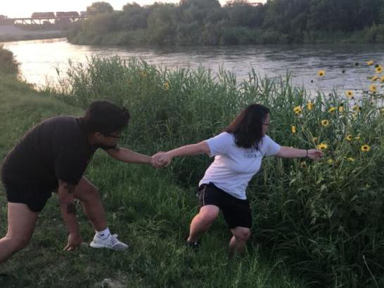 A man holding the hand of a woman to keep her from falling into a river as she reaches for a distant sunflower