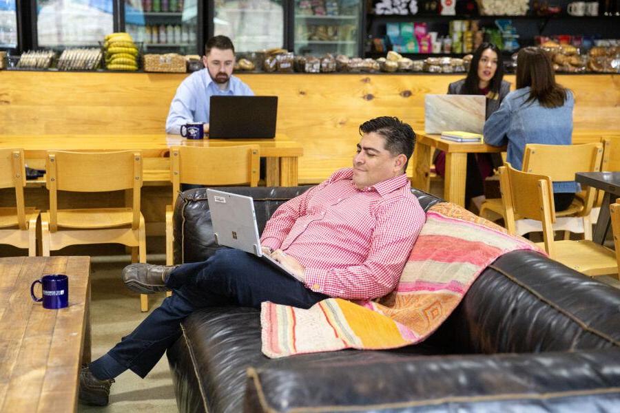 The image shows a relaxed setting in a café. A man in a red checkered shirt is sitting on a black leather sofa with a colorful blanket, 在笔记本电脑上工作. In the background, a man and two women are seated at tables, also using laptops. 咖啡馆有一个温暖的房间, inviting atmosphere with wooden furniture and shelves stocked with various food items. The focus is on a comfortable and productive environment where people are working and socializing.