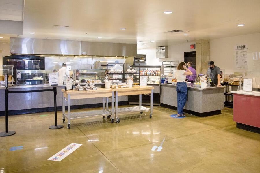 The image shows a spacious, modern cafeteria with various food stations. A few people are seen ordering and serving food, with one person in the foreground examining the options. The cafeteria has a clean and organized layout, featuring a selection of packaged foods, 饮料, 新鲜的饭菜. The floor is marked with social distancing indicators, and the atmosphere is calm and orderly. The setting suggests a focus on safety, efficiency, and a variety of dining choices.